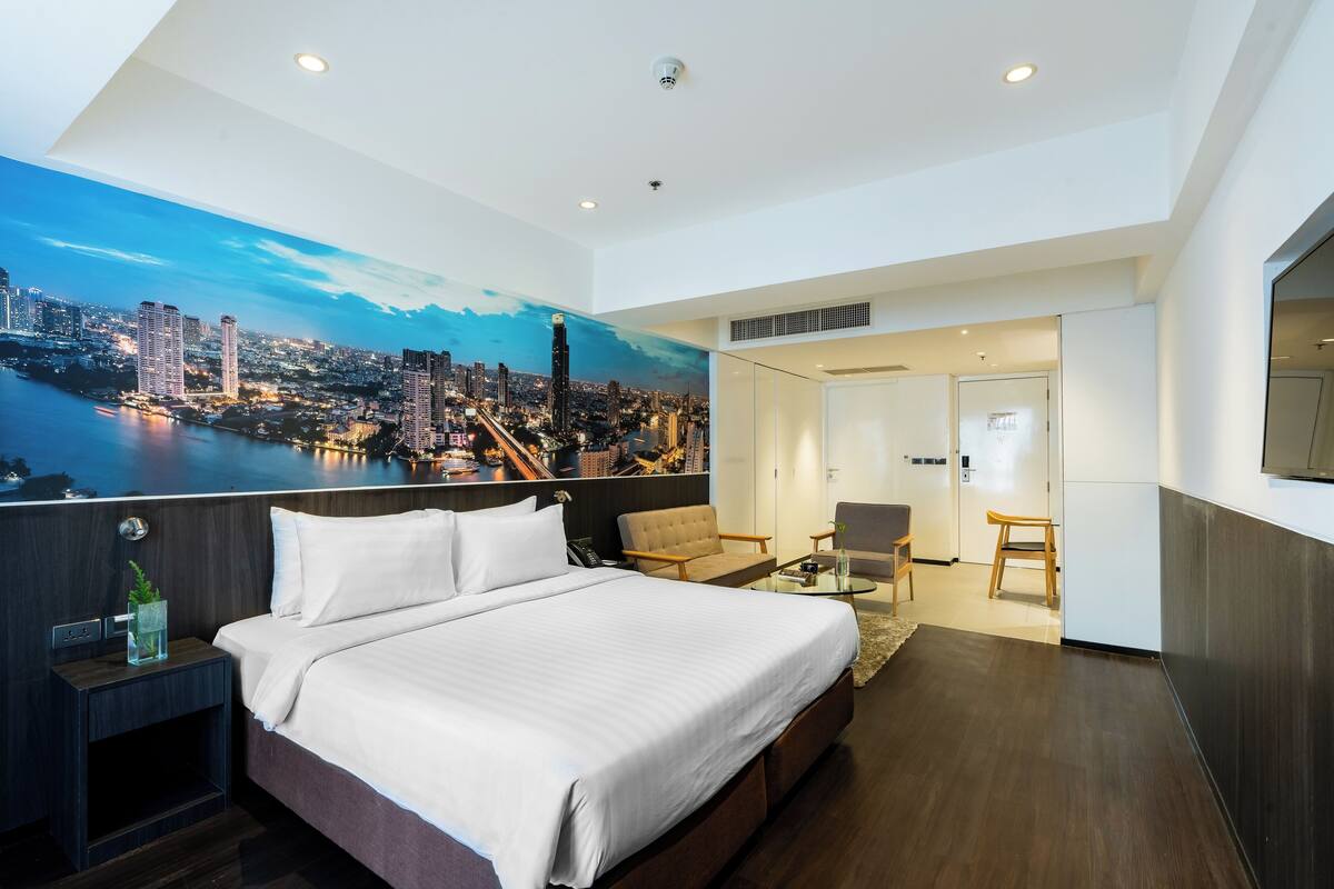 A modern hotel room with a large bed, a seating area, and a cityscape mural on the wall.