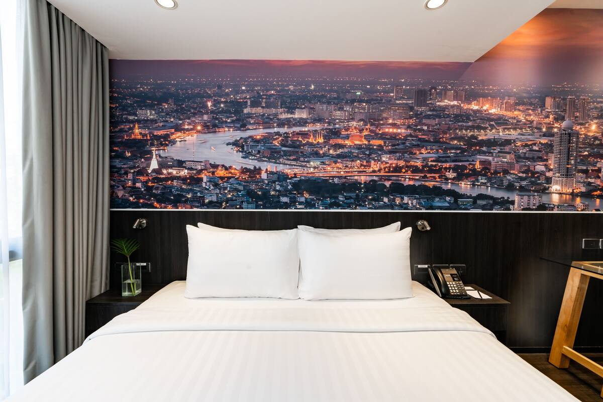 A hotel room with a large bed, white bedding, a nightstand with a phone, and a view of a cityscape at dusk.