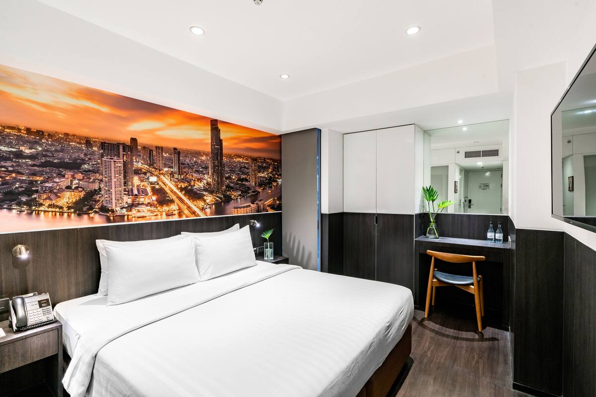 A hotel room with a large bed, a desk with a chair, a mirror, and a cityscape mural on the wall.