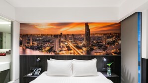 Une chambre d’hôtel avec une grande fresque représentant un paysage urbain sur le mur, un lit avec du linge de maison blanc, une table de chevet avec un téléphone et une petite plante.