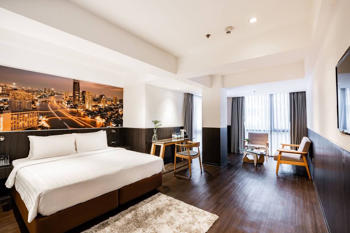 A modern hotel room with a large bed, a desk, two chairs, and a cityscape view.