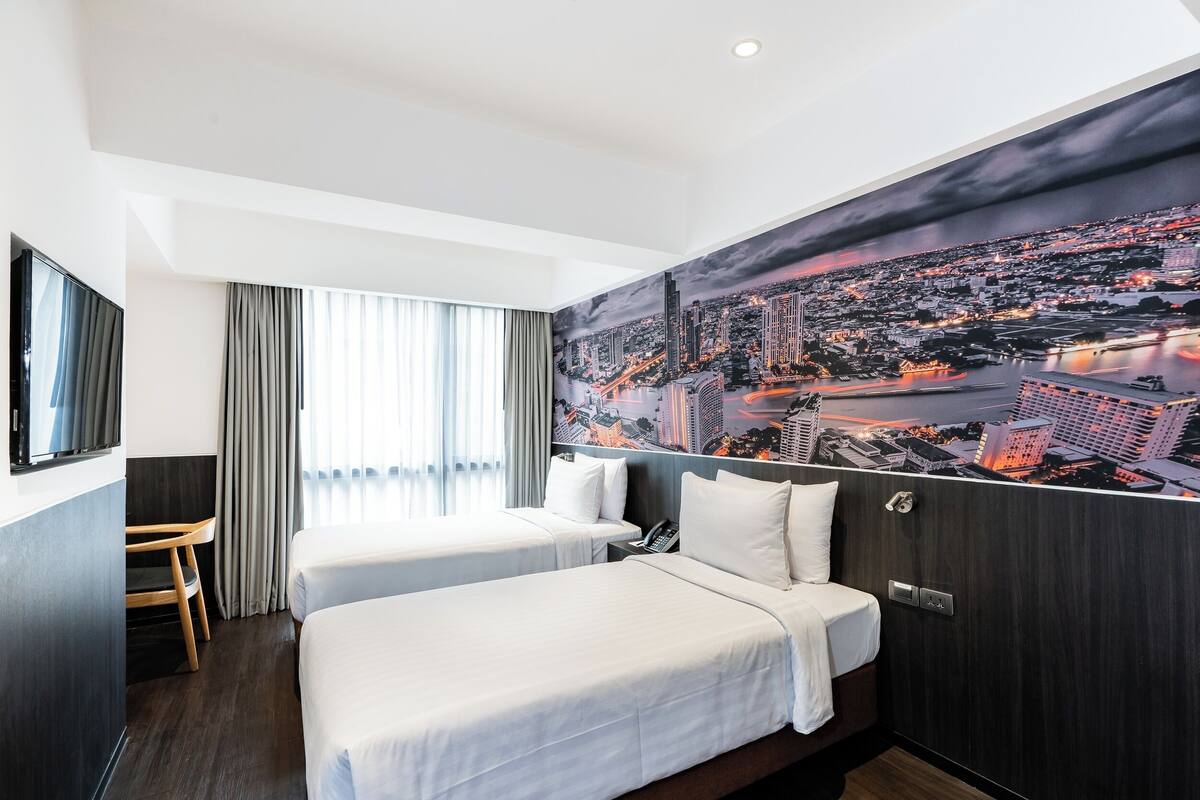 A hotel room with two beds, a desk, a chair, and a large cityscape mural on the wall.