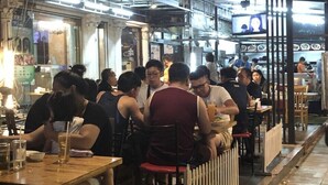 A crowded outdoor dining area with tables and chairs, a white picket fence, and a building with a sign in the background. - At Bangkok Hotel (Bangkok)