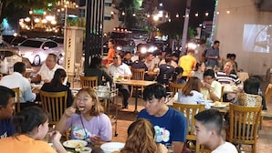 A crowded outdoor dining area with wooden tables and chairs, surrounded by buildings and street lights. - At Bangkok Hotel (Bangkok)