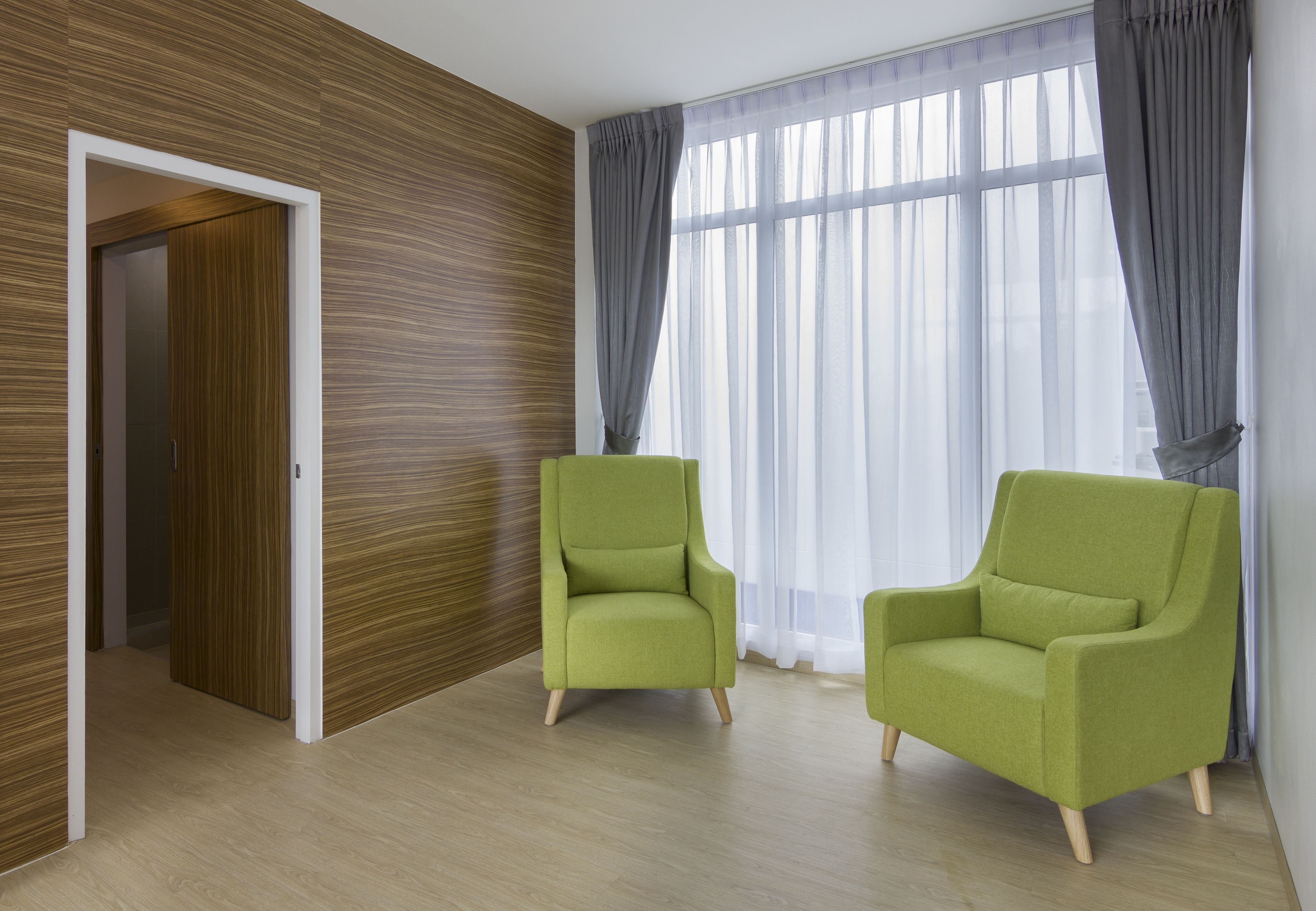 A room with wooden panel walls, a large window with sheer curtains, and two green armchairs.
