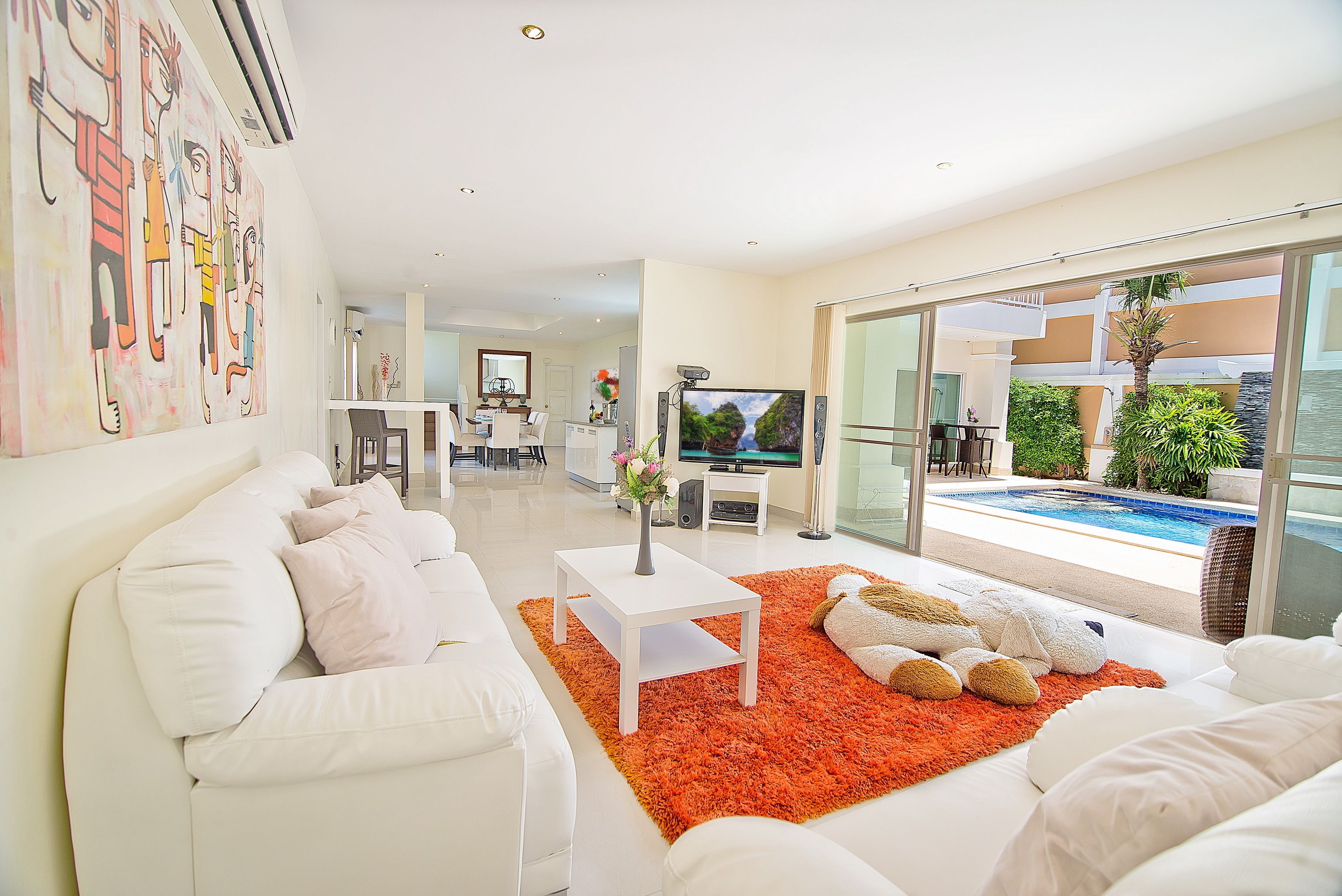 Uma sala de estar moderna com um sofá branco, uma mesa de centro e uma TV. Dá para ver uma piscina através das portas de vidro.