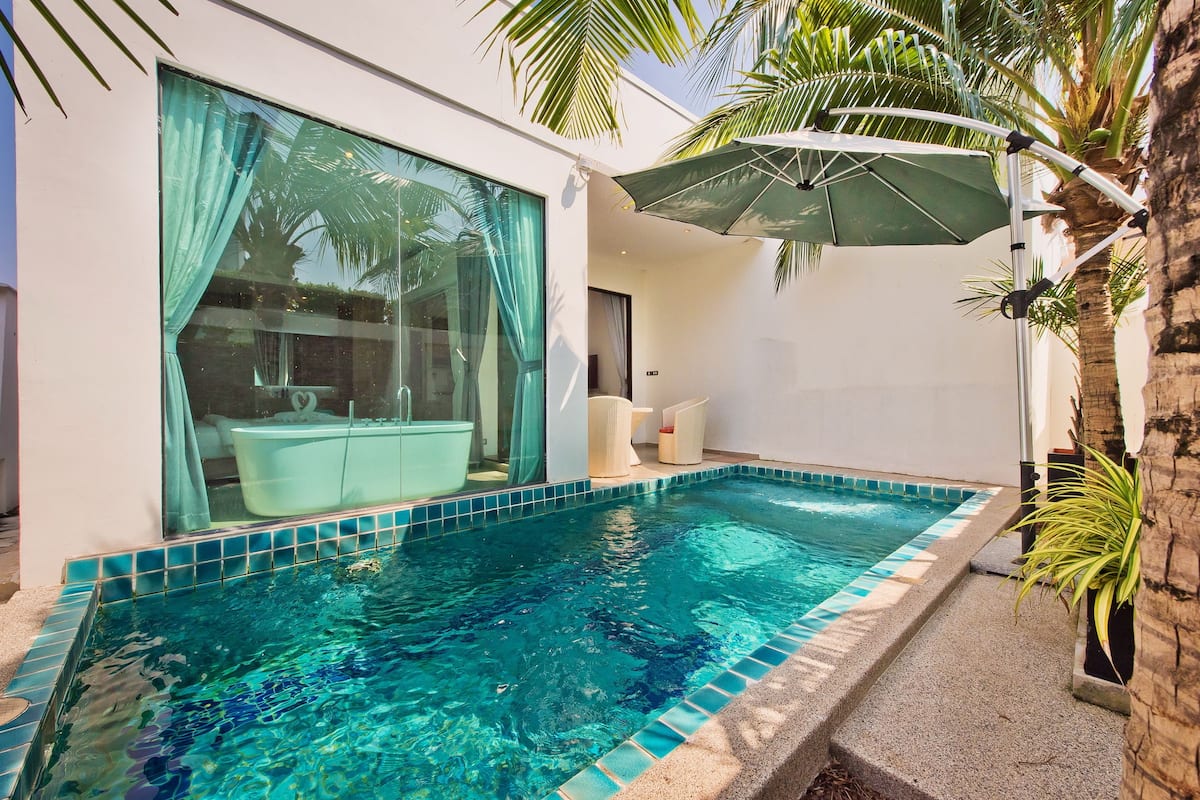 Un área de piscina moderna con una piscina azul cristalina, una puerta de vidrio con cortinas y una silla blanca bajo un paraguas verde.