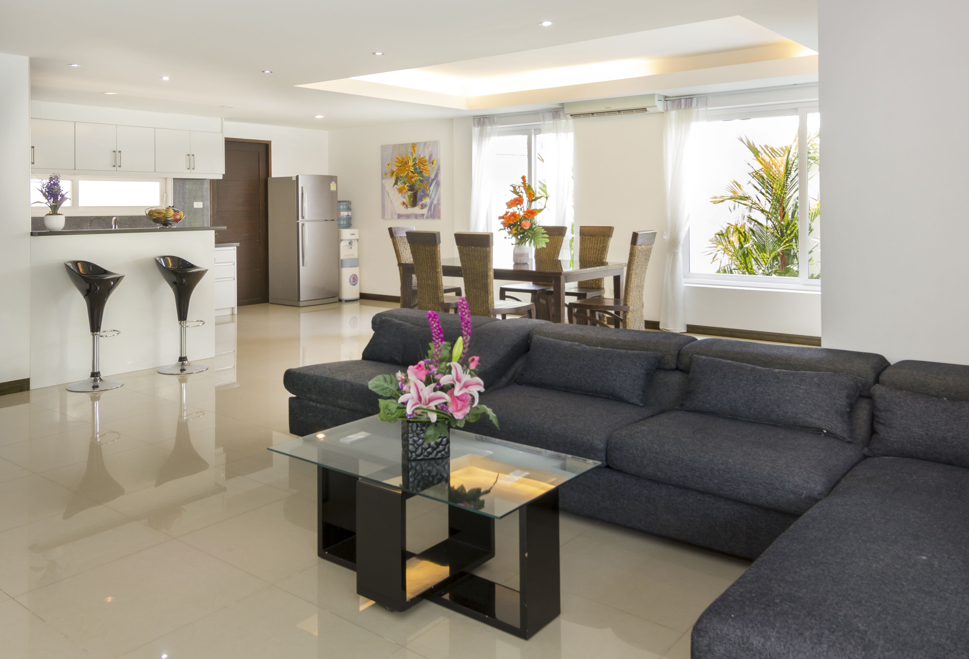 Un salon moderne avec un canapé gris foncé, une table basse en verre ornée d’un vase de fleurs, un coin cuisine avec des meubles blancs et un coin repas avec une table et des chaises.