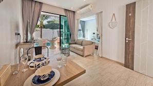A modern living room with a sofa, dining table, and a glass door leading to a balcony. - Tropicana Pool Villa (Pattaya)