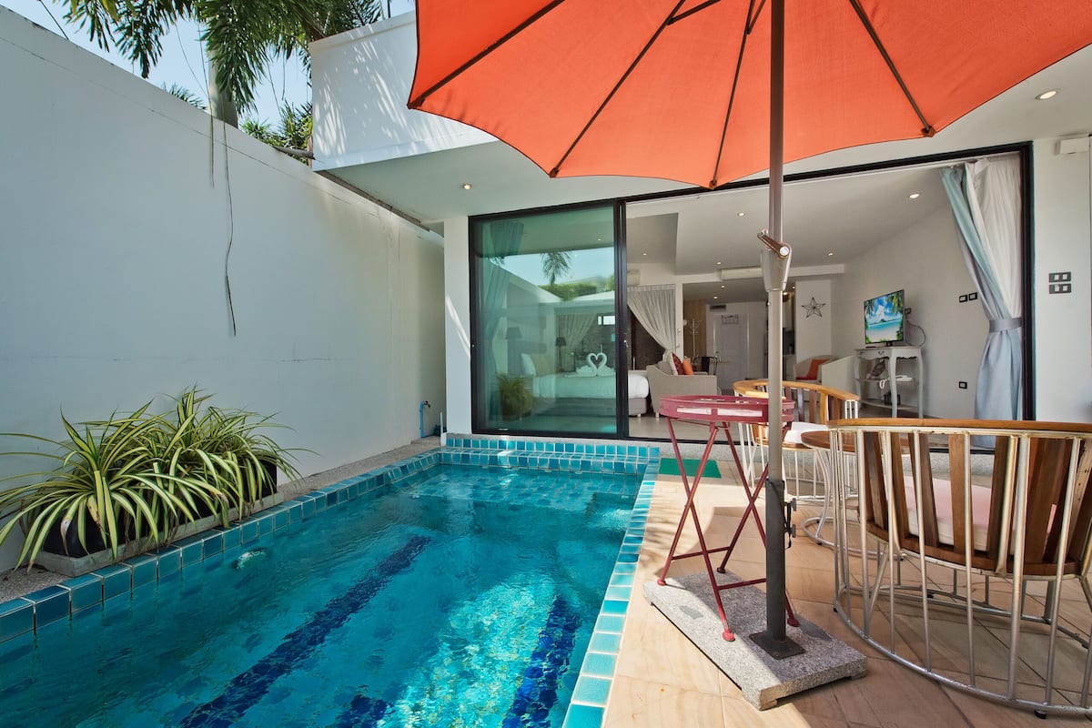 Una piscina con borde de azulejos, rodeada de plantas en macetas, una terraza de madera y un ventanal que da a un espacio interior con una mesa y sillas de comedor.