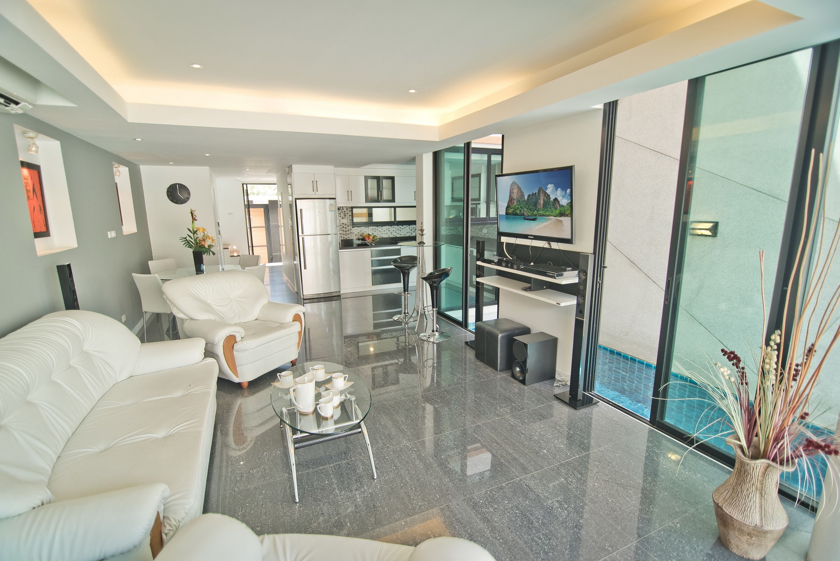 A modern living room with a sofa, coffee table, and a TV. There is a kitchen area with cabinets and a refrigerator. A glass door leads to a pool area.