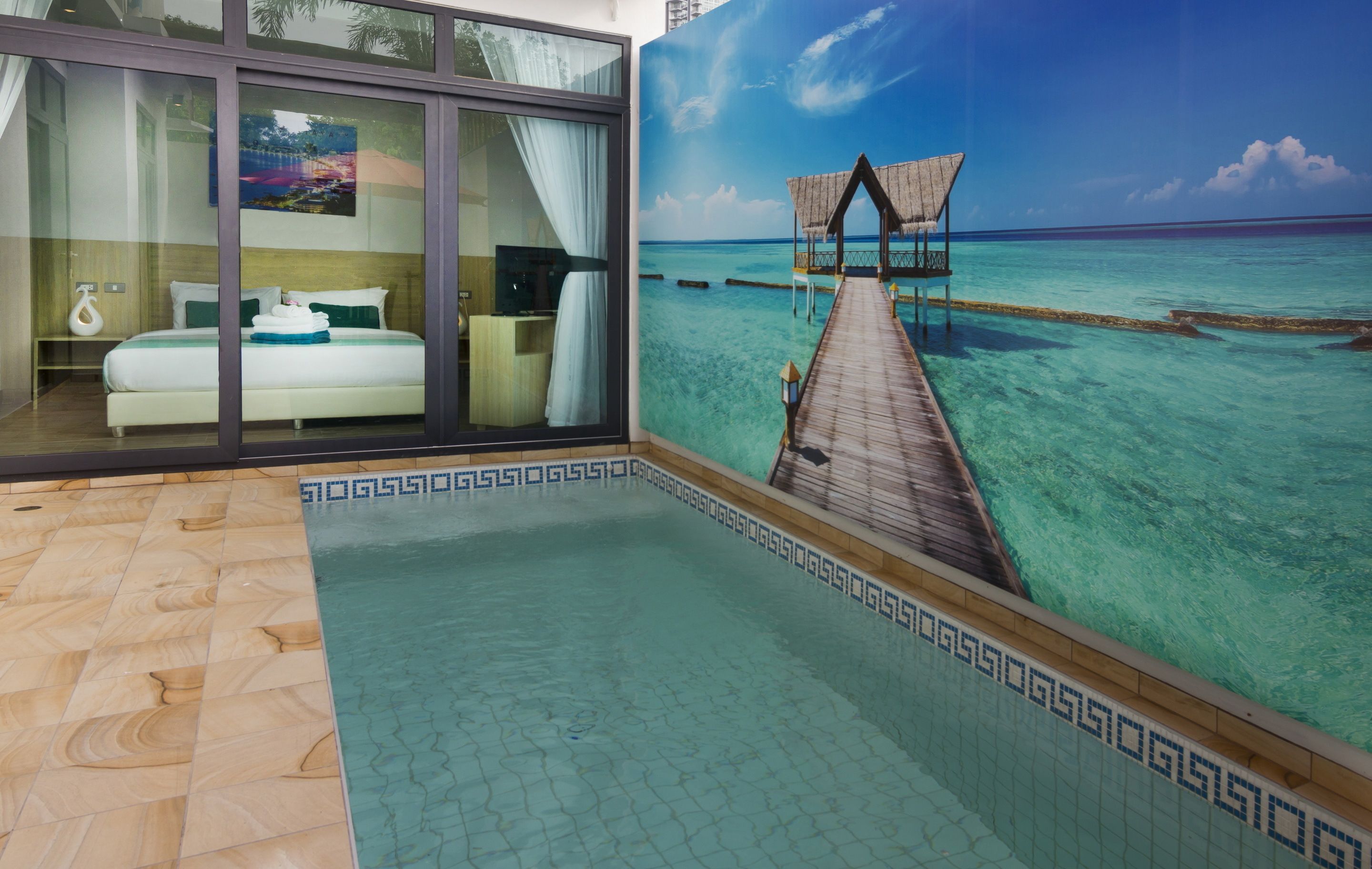 A hotel room with a large window, a bed, a nightstand, and a view of a wooden pier leading to overwater bungalows.