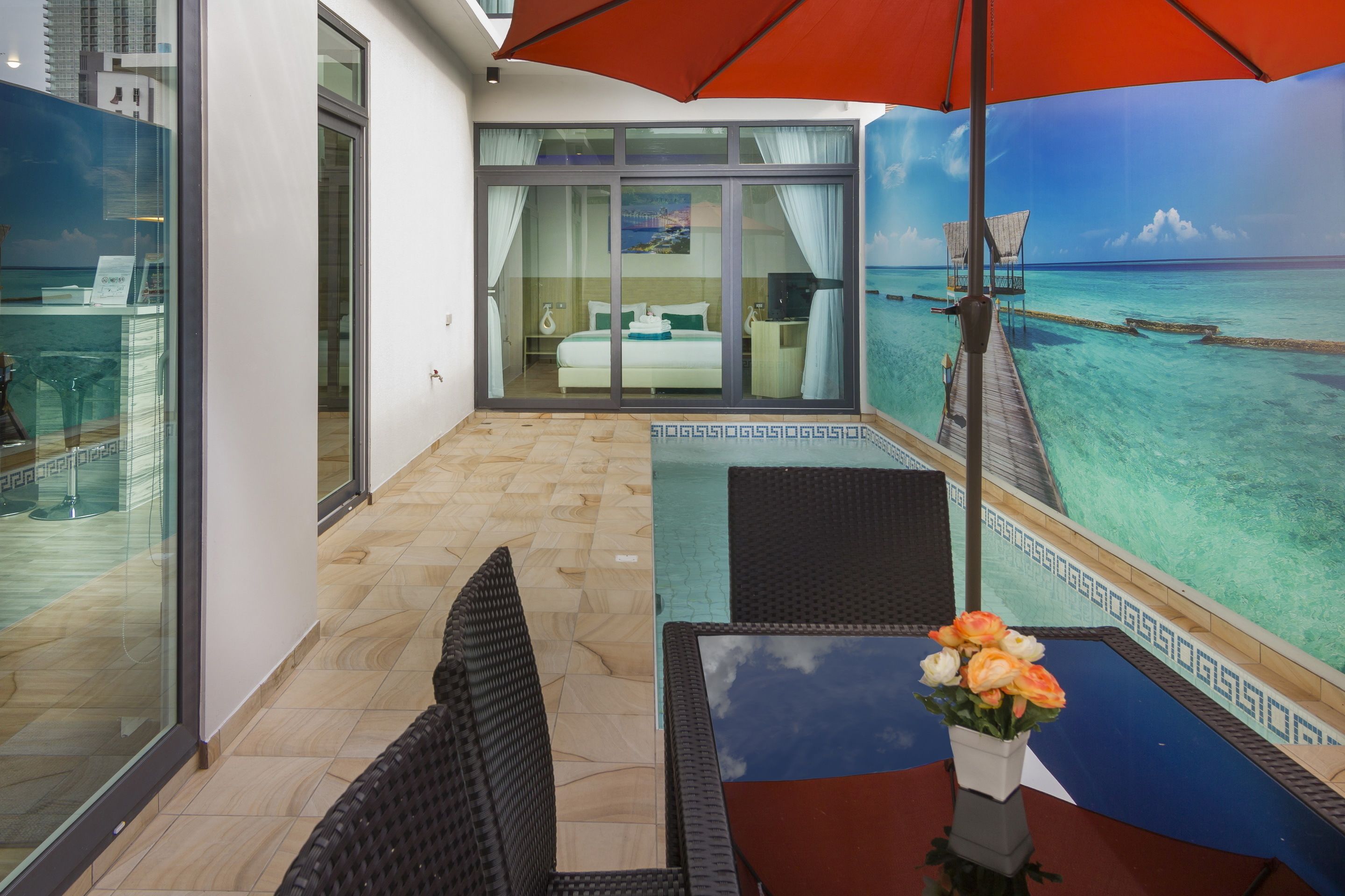 A balcony with a glass door leading to a bedroom, a pool area with a table and chairs, and a view of the ocean.