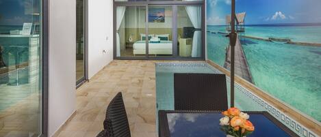 A balcony with a glass door leading to a bedroom, a pool area with a table and chairs, and a view of the ocean.
