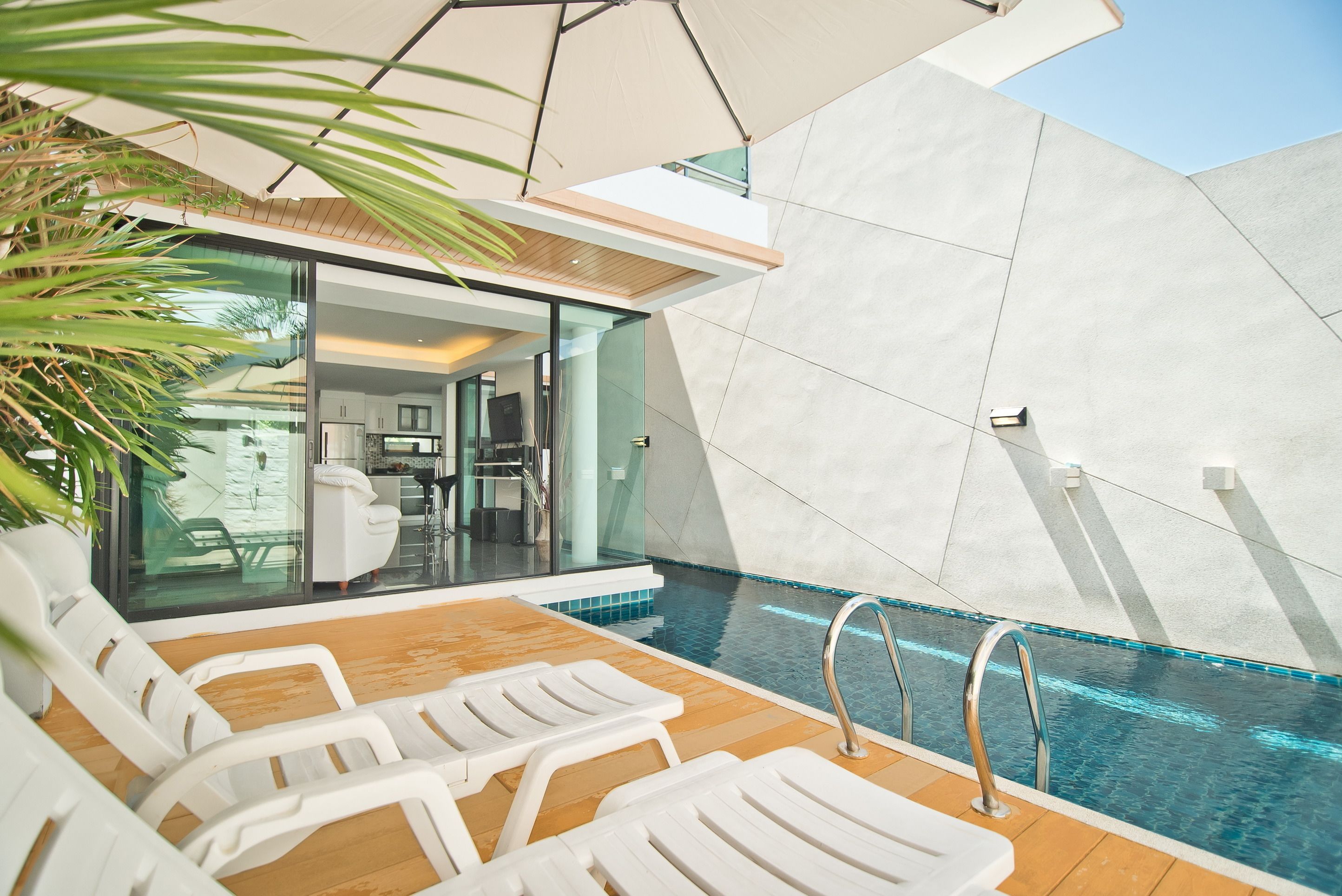 A modern outdoor pool area with white lounge chairs, a wooden deck, and a glass-enclosed kitchen visible through the glass walls.