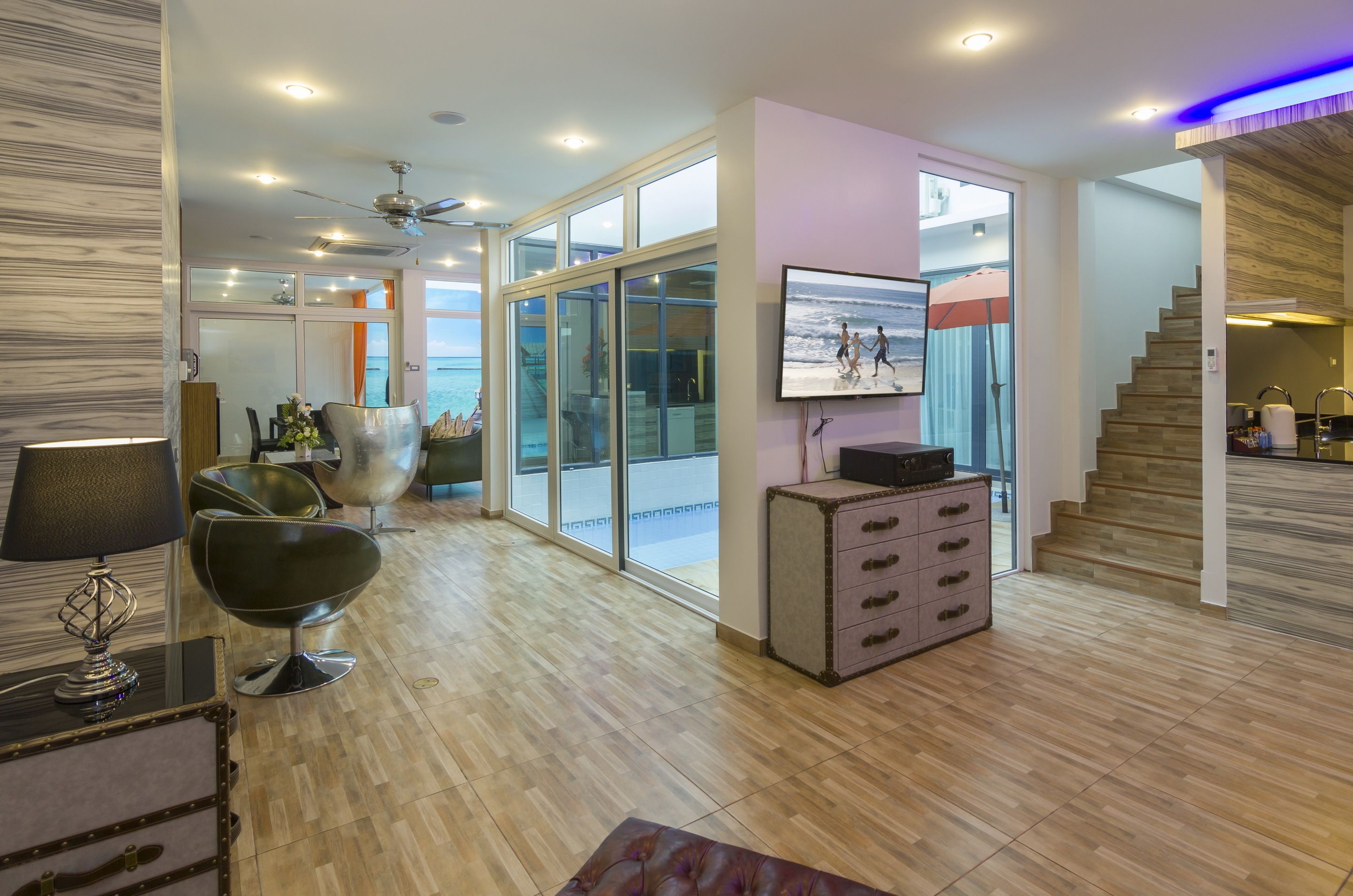 A modern living room with a large glass door leading to a balcony, a flat-screen TV mounted on the wall, and a staircase to the upper floor.