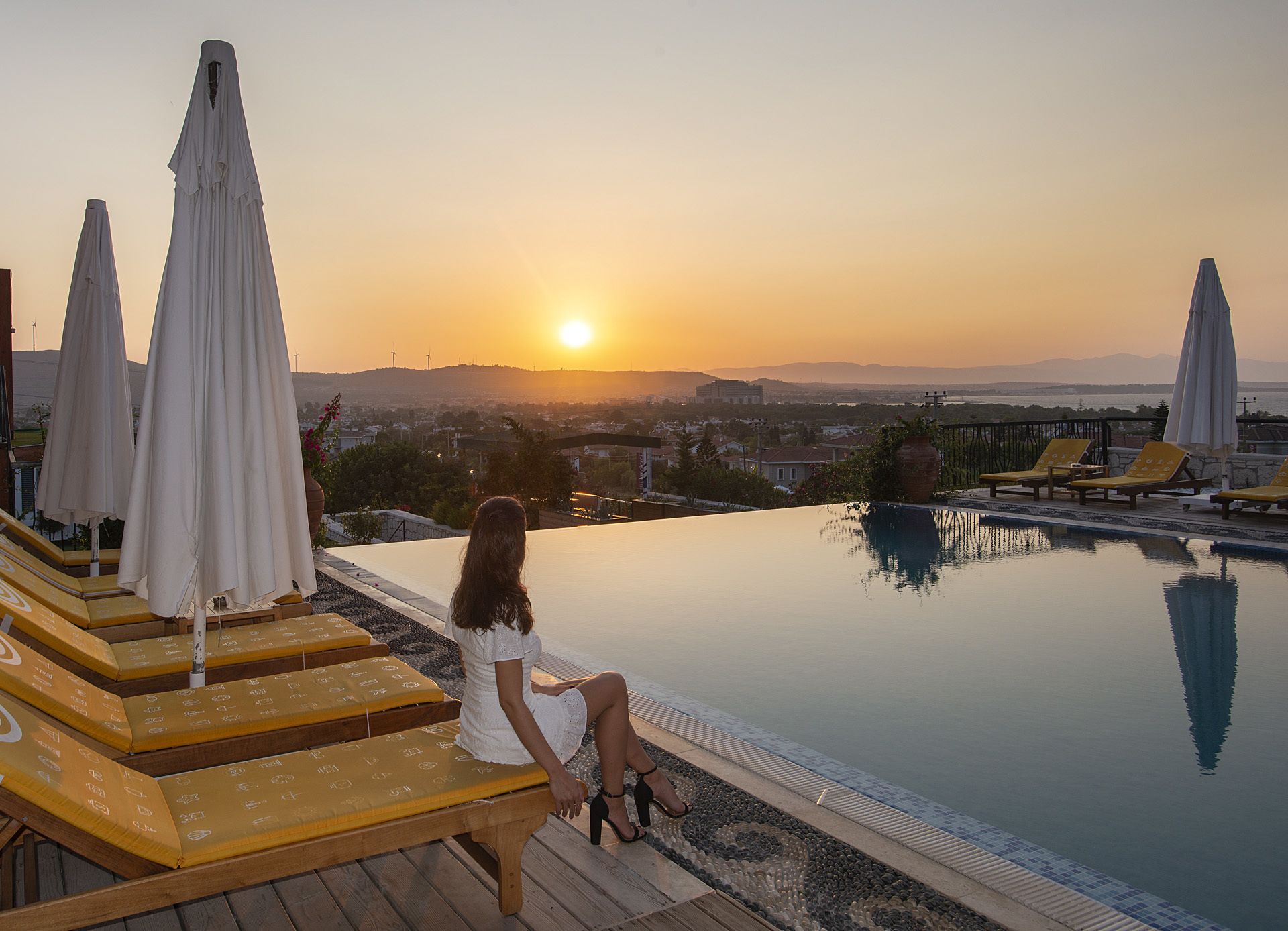 A woman sitting by a pool with a sunset view, lounge chairs, and umbrellas.