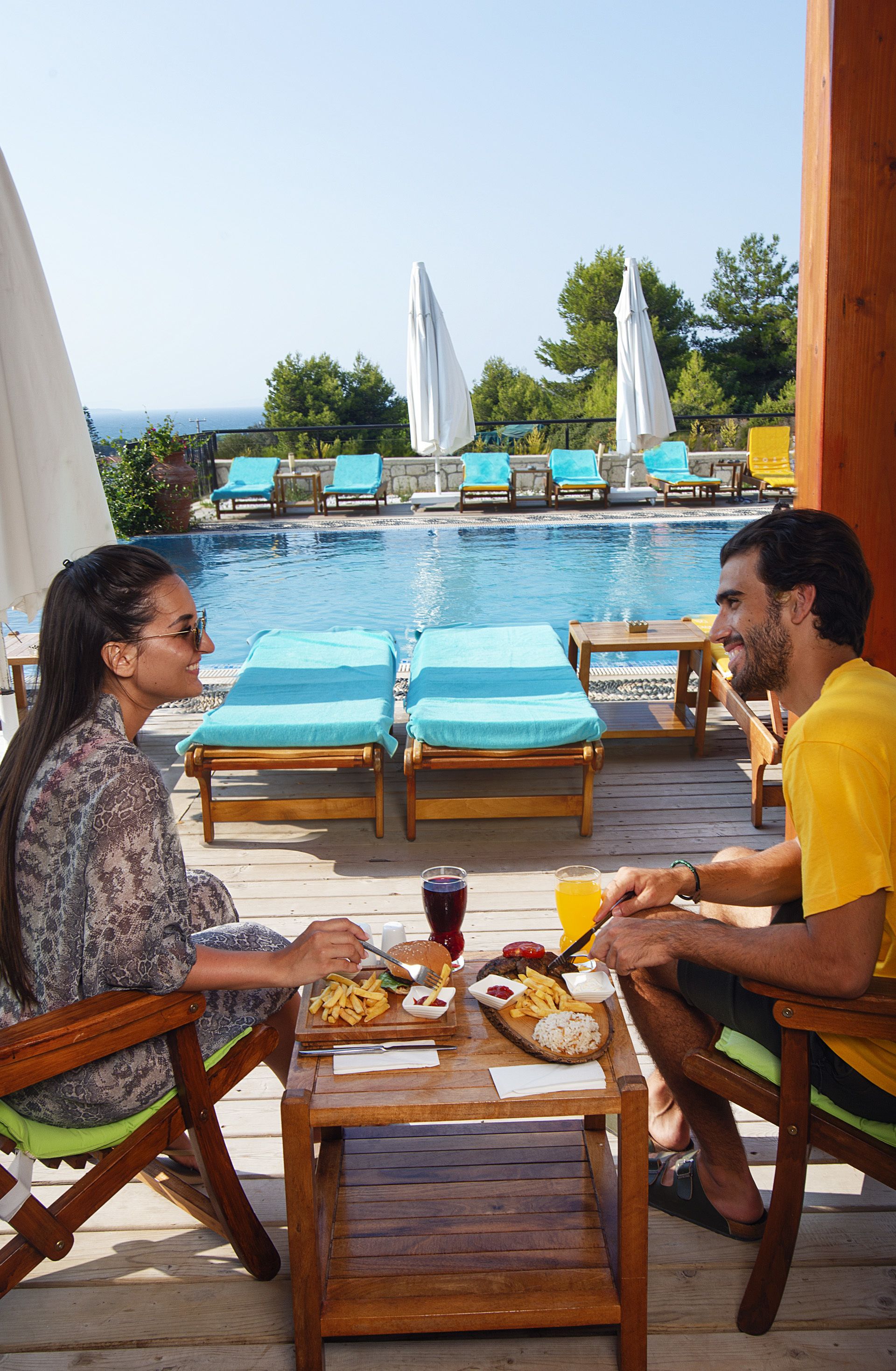 A couple dining at a wooden table by a poolside with lounge chairs and umbrellas in the background.