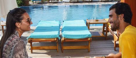 A couple dining at a wooden table by a poolside with lounge chairs and umbrellas in the background.
