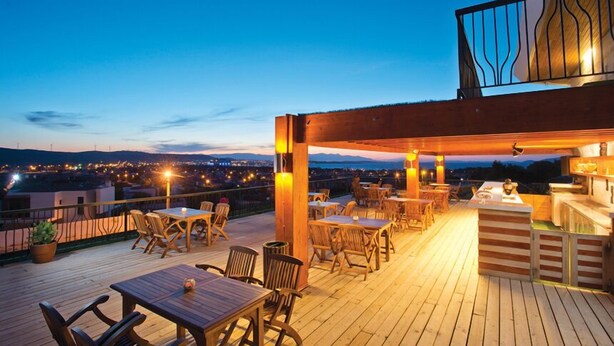 A wooden deck with tables and chairs, a bar area, and a view of the cityscape at dusk. - Alacati Villa Renk (Cesme)