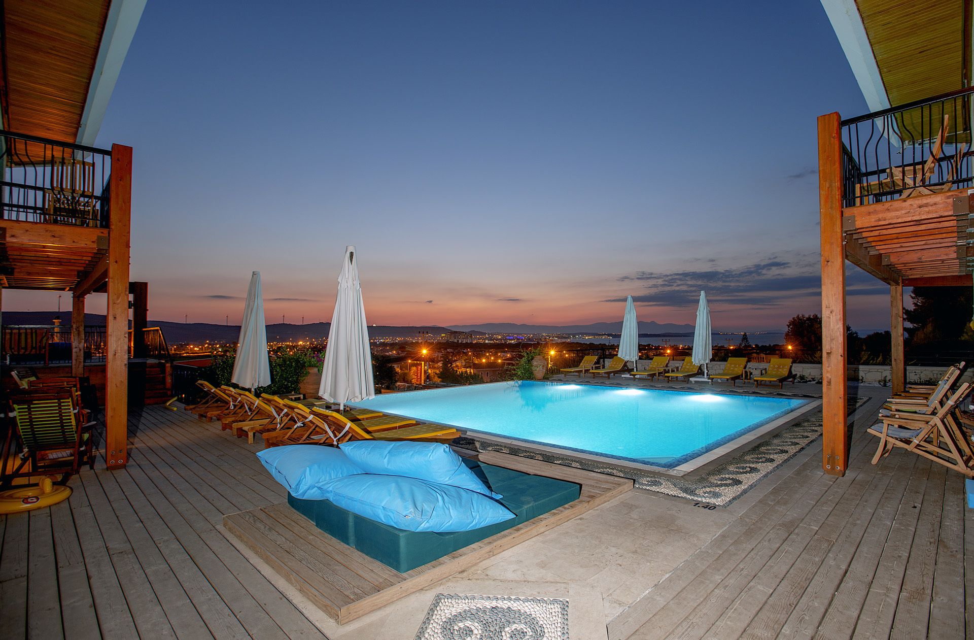 Ein Poolbereich auf dem Dach mit Liegestühlen und Sonnenschirmen mit Blick auf eine Stadtlandschaft in der Abenddämmerung.