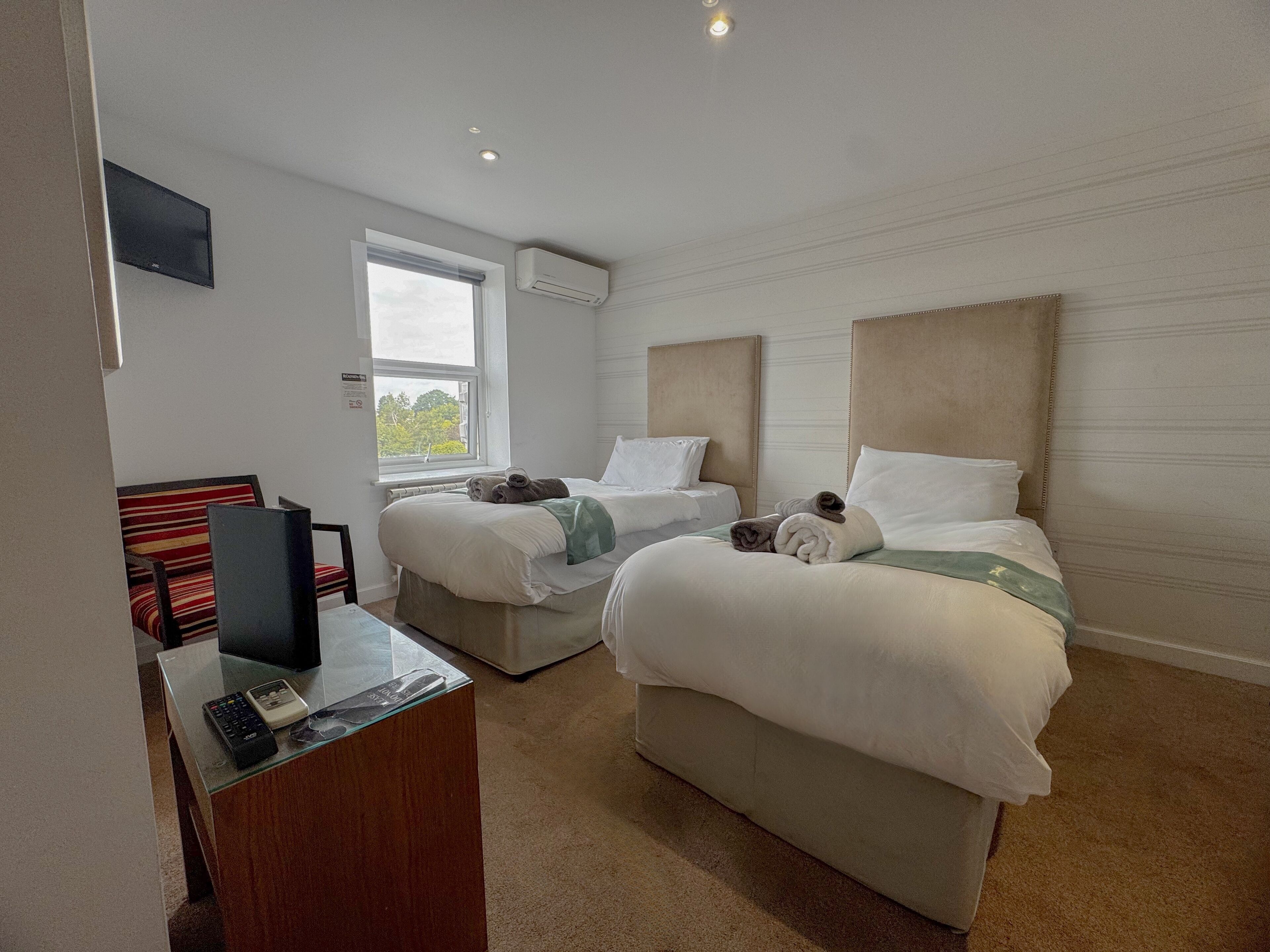 A hotel room with two beds, a desk with a computer, and a window with a view of trees.