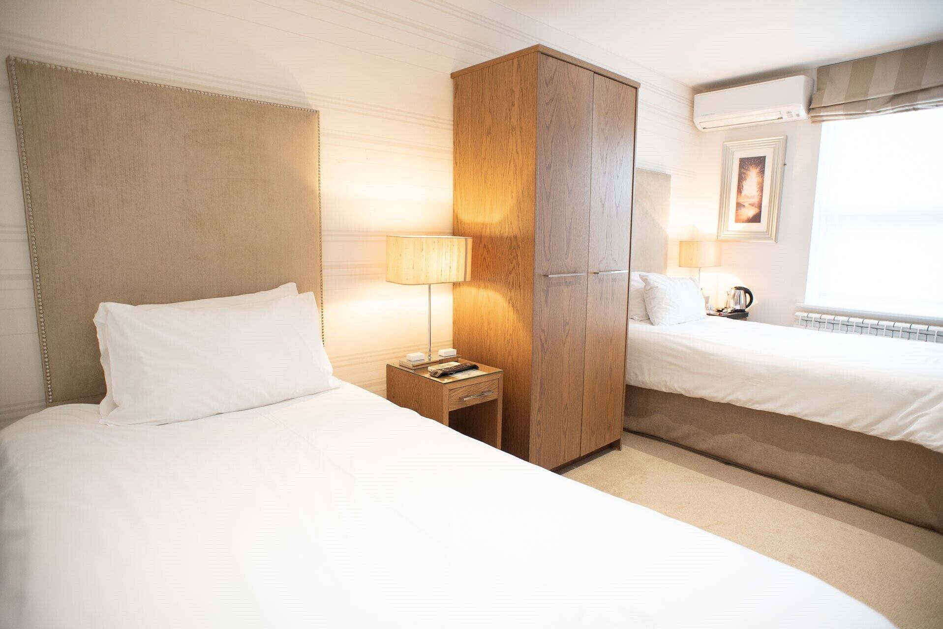 Une chambre d’hôtel avec deux lits, une armoire en bois, une table de chevet avec une lampe et une fenêtre avec des rideaux.