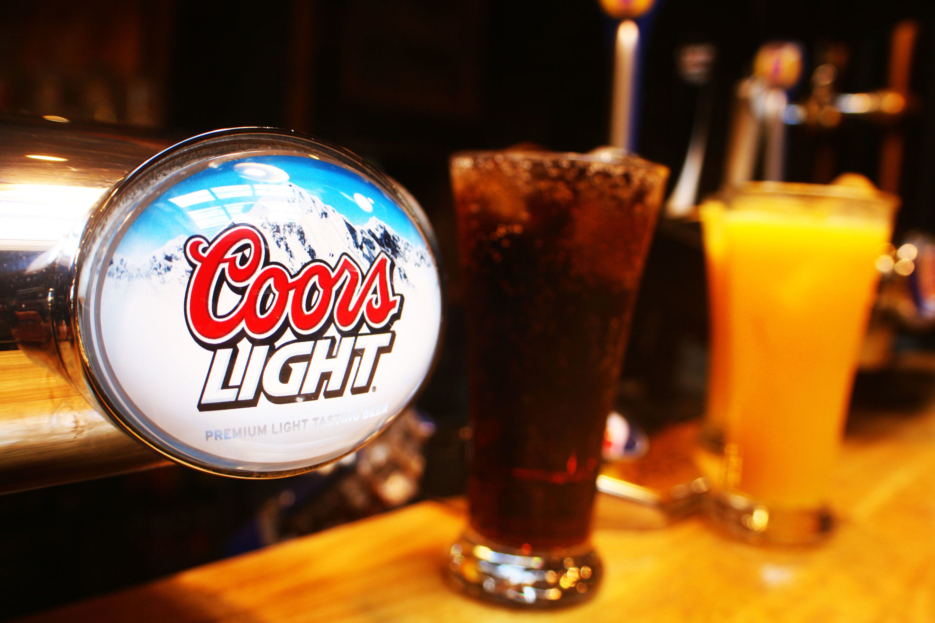 Een Coors Light bierfles met etiket, een glas donker bier en een glas sinaasappelsap op een barkruk.