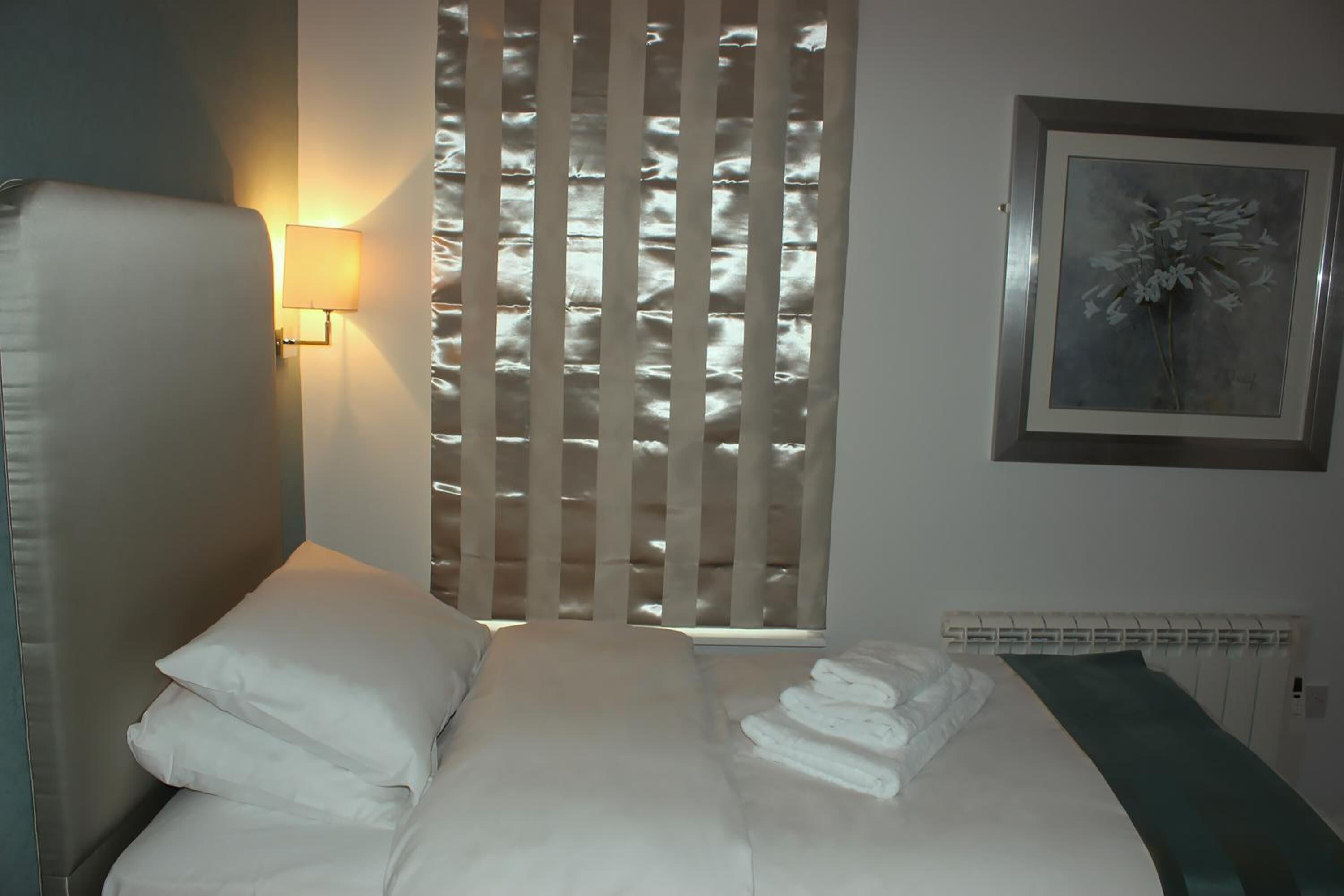 A neatly made bed with white linens, a wall-mounted lamp, and a framed artwork on the wall.