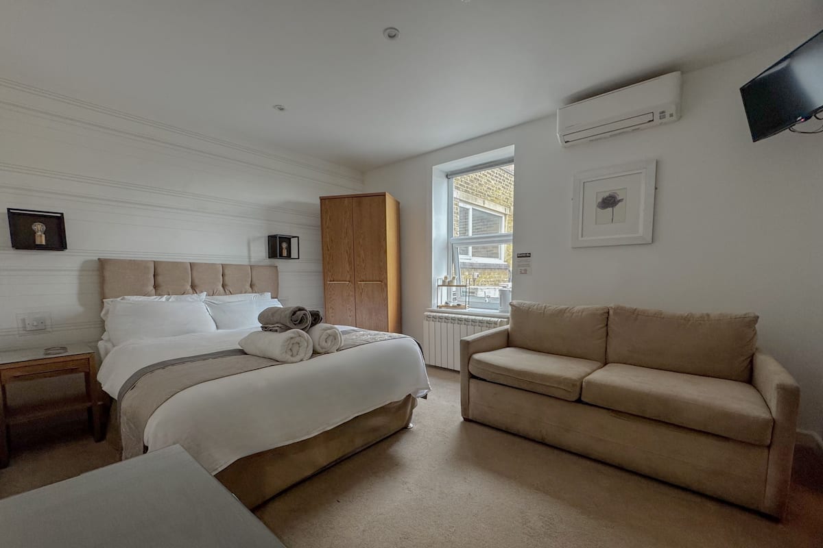 A neatly arranged bedroom with a large bed, a wooden wardrobe, a beige sofa, and a small table.