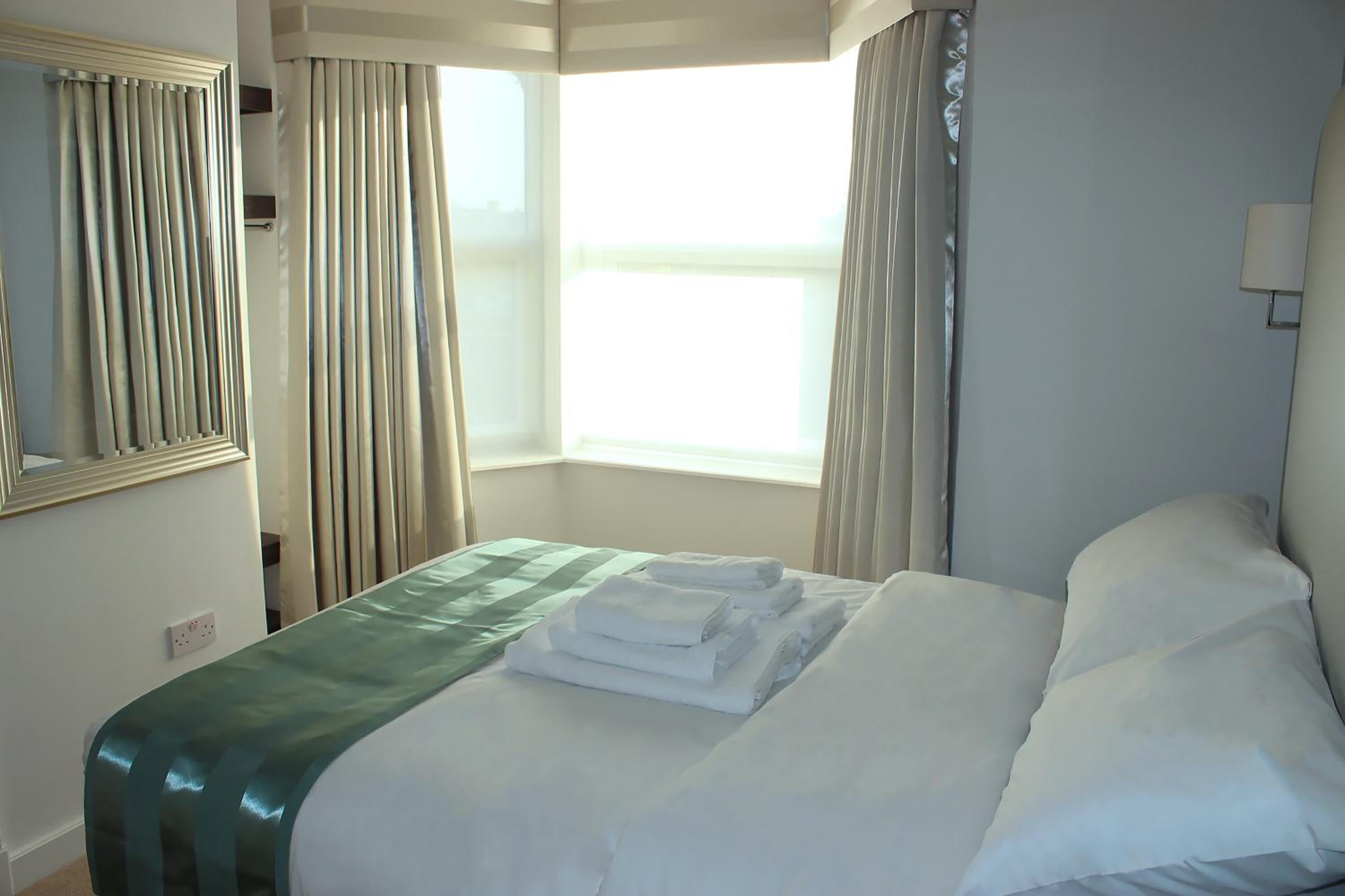 A hotel room with a bed, a window with curtains, a mirror, and a wall-mounted lamp.