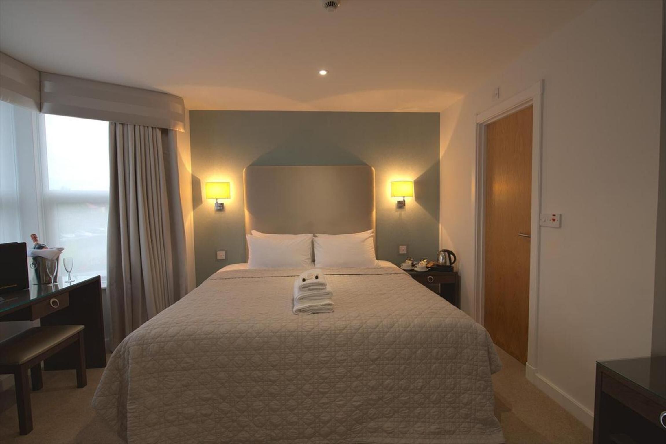 A hotel room with a large bed, two bedside lamps, a desk with a computer, and a window with curtains.