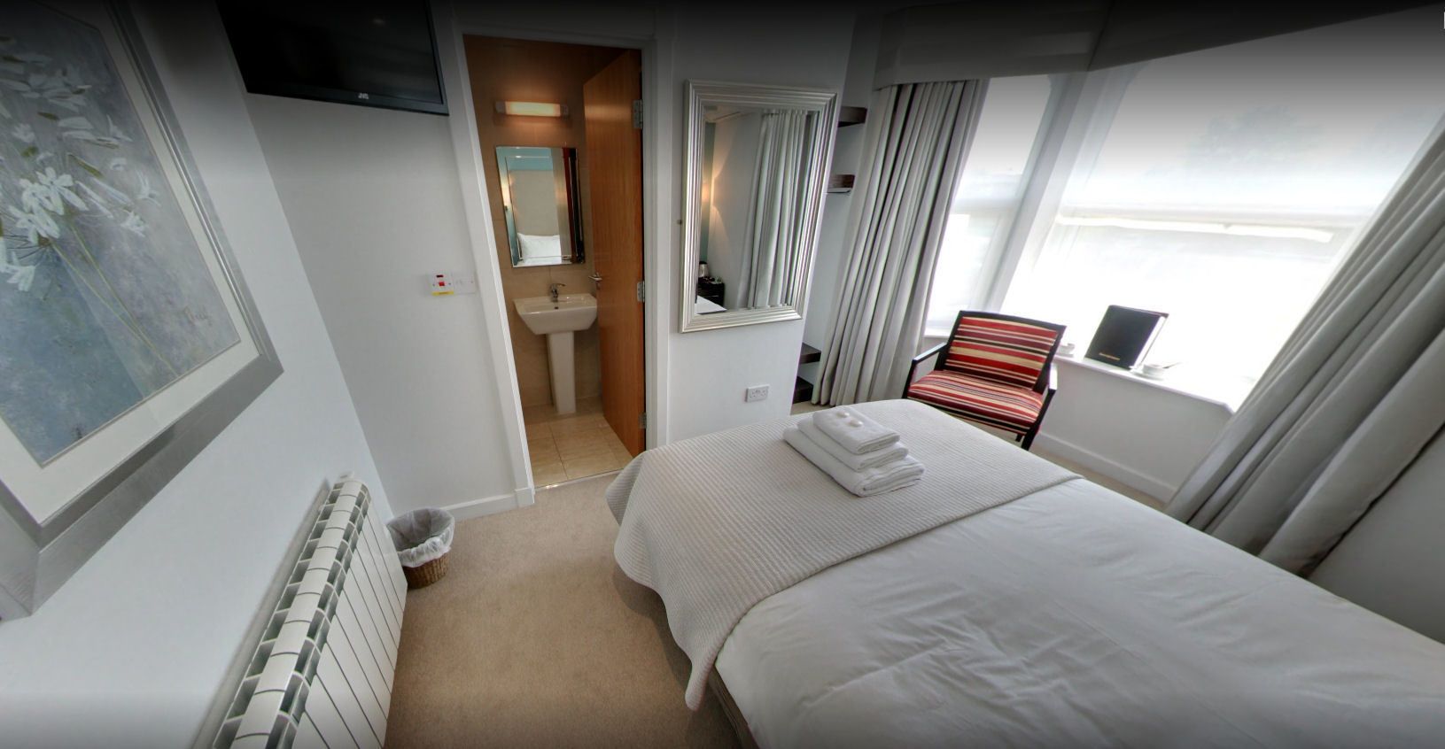 A hotel room with a bed, a window with curtains, a bathroom with a sink and mirror, and a radiator.