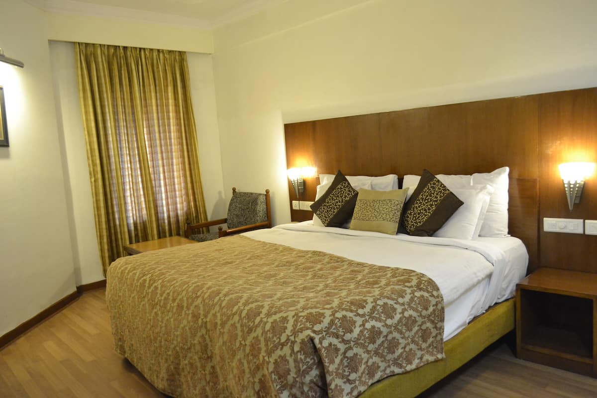 Habitación de hotel con una cama grande, un cabecero de madera, dos mesitas de noche, una silla y una ventana con cortinas.