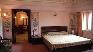 A bedroom with a large bed, a nightstand, a chair, a desk, and a door leading to another room. - Hotel Jaipur Ashok (Jaipur)