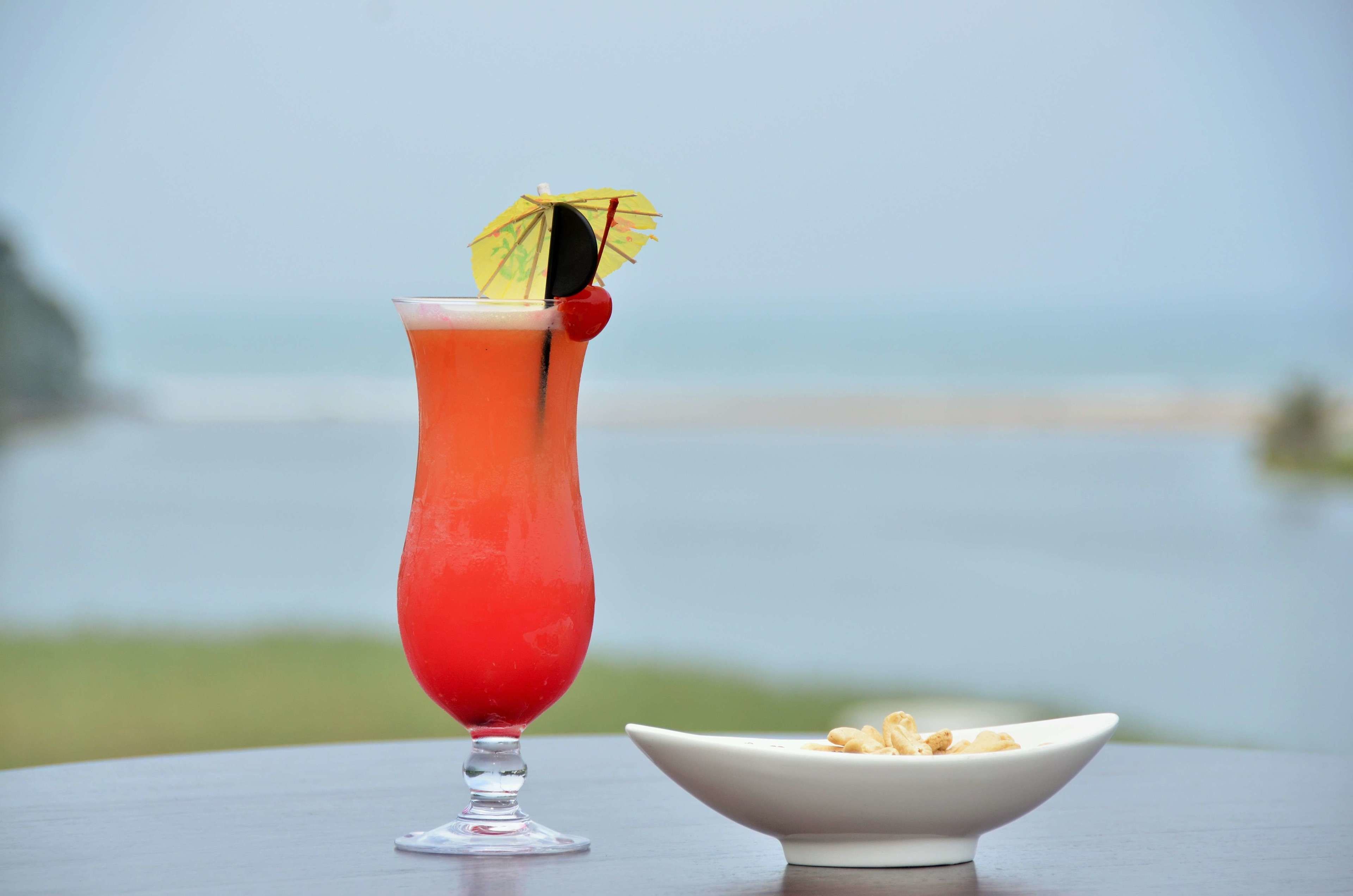 A tall glass of red drink with a cherry and a yellow umbrella on a table with a view of the ocean.