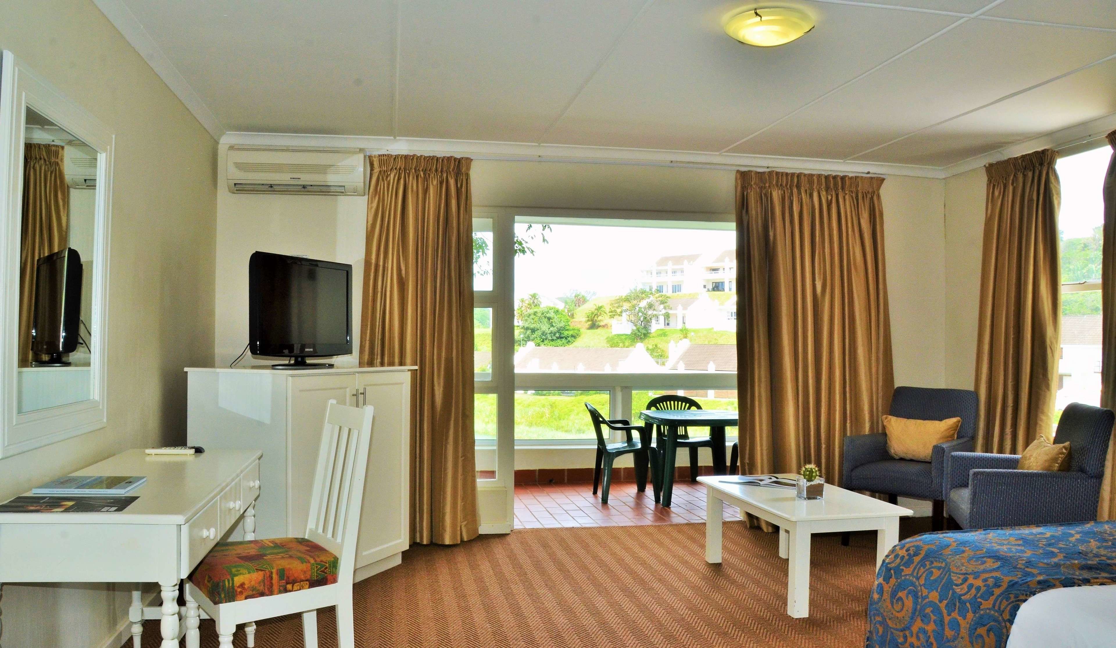 A hotel room with a television on a cabinet, a desk, a chair, a sofa, and a view of the outdoors.