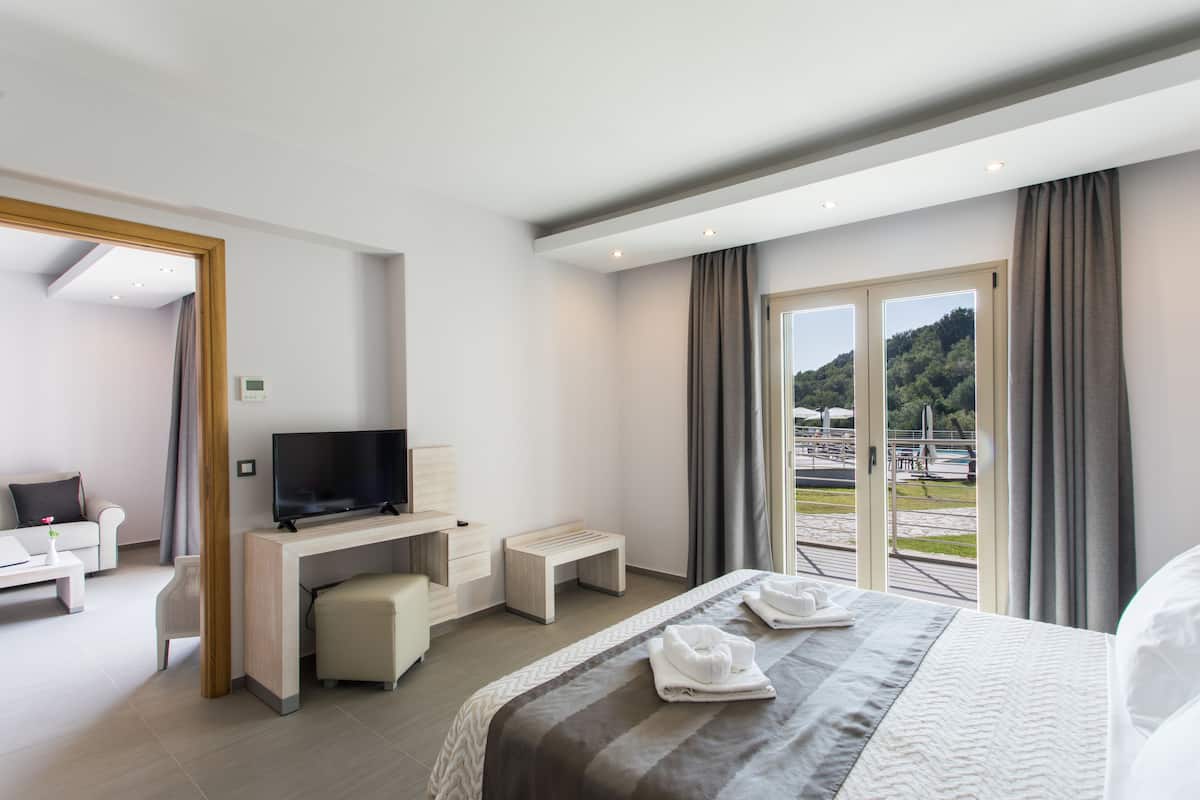 A modern hotel room with a bed, a TV, a desk, and a balcony with a view of a forest.