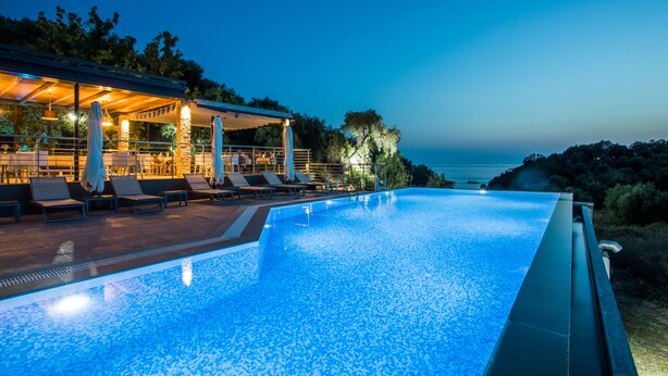 A swimming pool with lounge chairs and a covered area, overlooking a body of water at dusk. - Aqua Oliva Resort (Igoumenitsa)