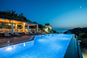 Piscina con sillas de descanso y un área cubierta, con vista a una extensión de agua al atardecer.