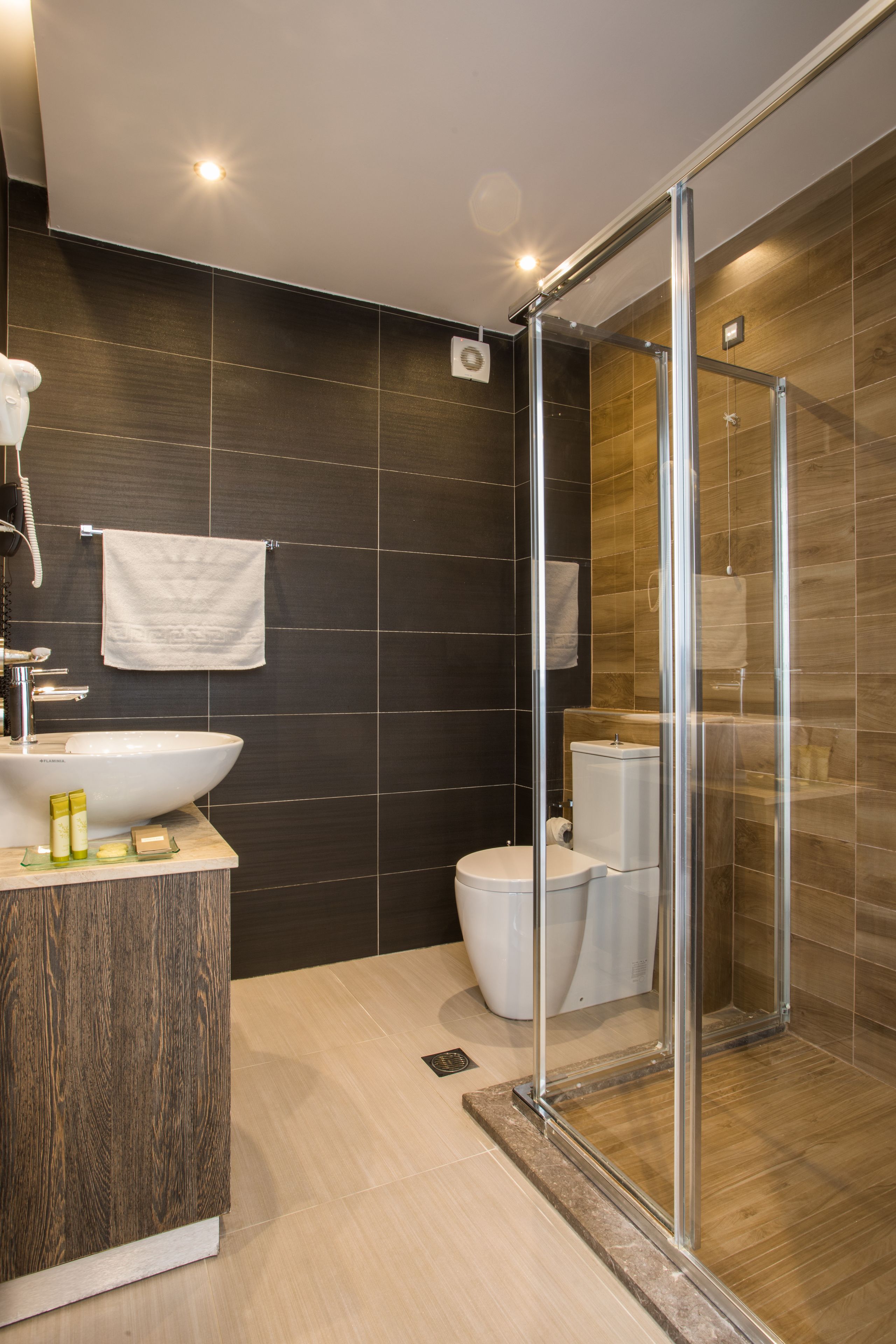A modern bathroom with a glass-enclosed shower, a wooden vanity, and a dark tiled wall.