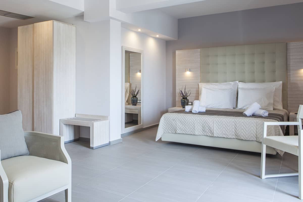 A modern hotel room with a large bed, two white chairs, a small table, and a mirror.