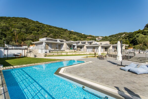 Una piscina con agua cristalina azul, rodeada por una zona pavimentada con sillas de descanso y sombrillas. Se observan edificios de diseño moderno al fondo, y un paisaje montañoso con vegetación.