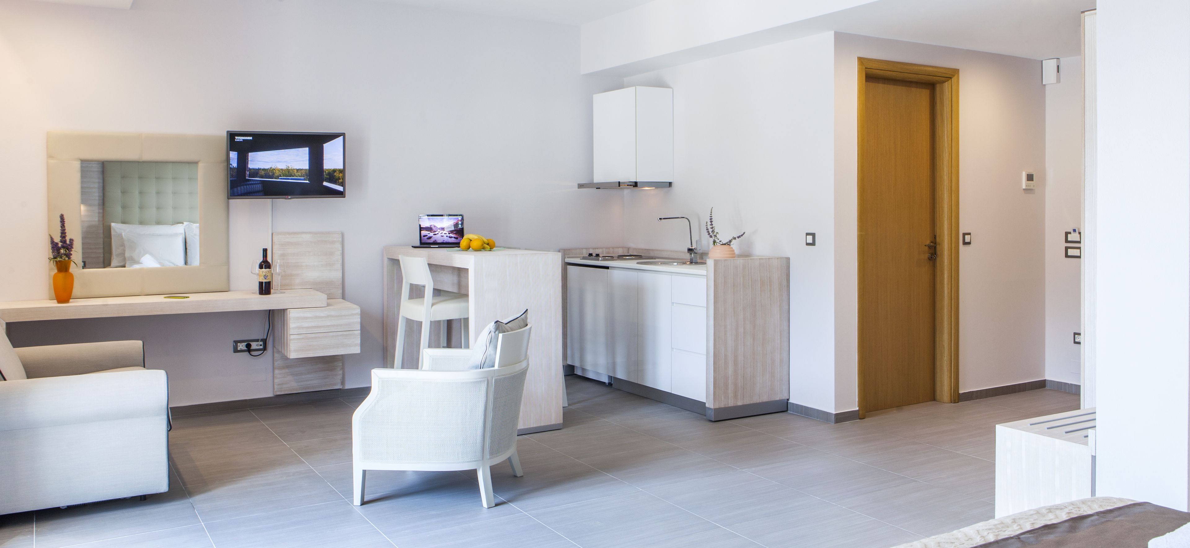 A modern living room with a sofa, a TV mounted on the wall, a small dining area with a table and chairs, and a kitchen with cabinets and a sink.