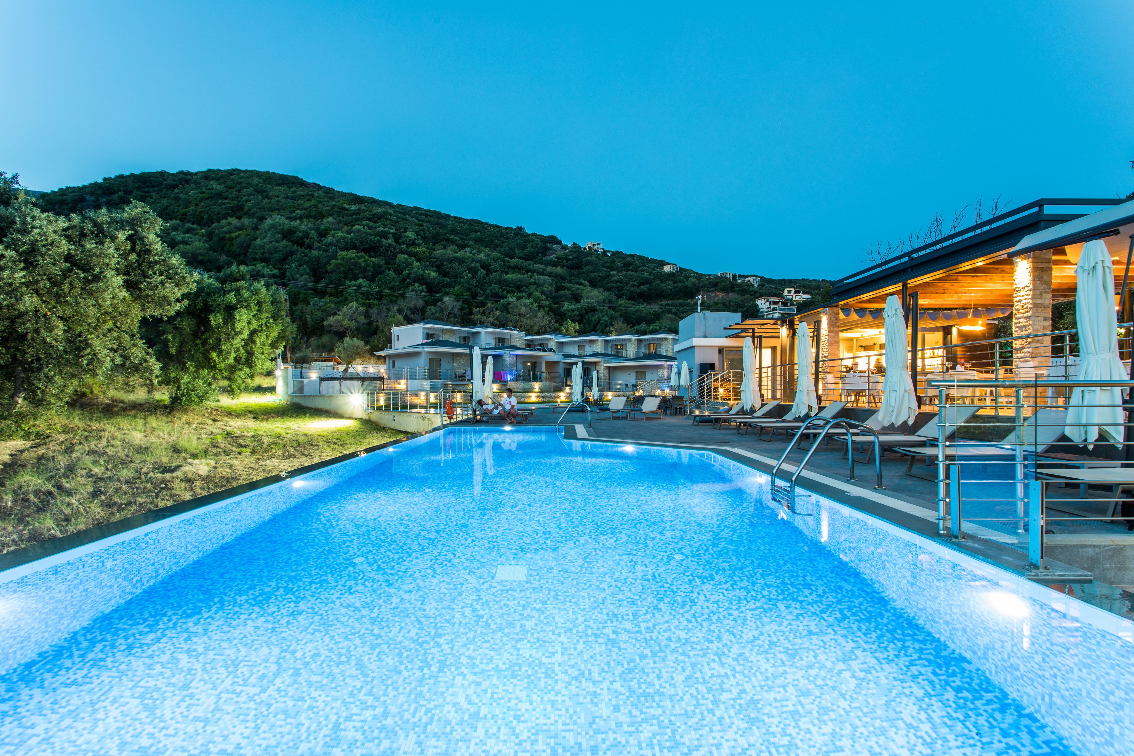 A swimming pool with lounge chairs and umbrellas, surrounded by buildings and a hillside.