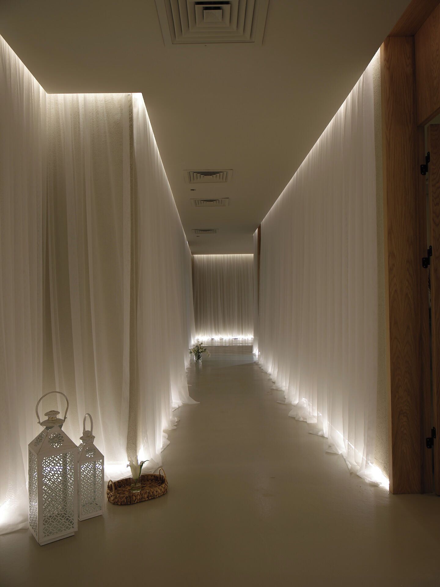 A corridor with sheer white drapes, a wooden door, and two lanterns on the floor.