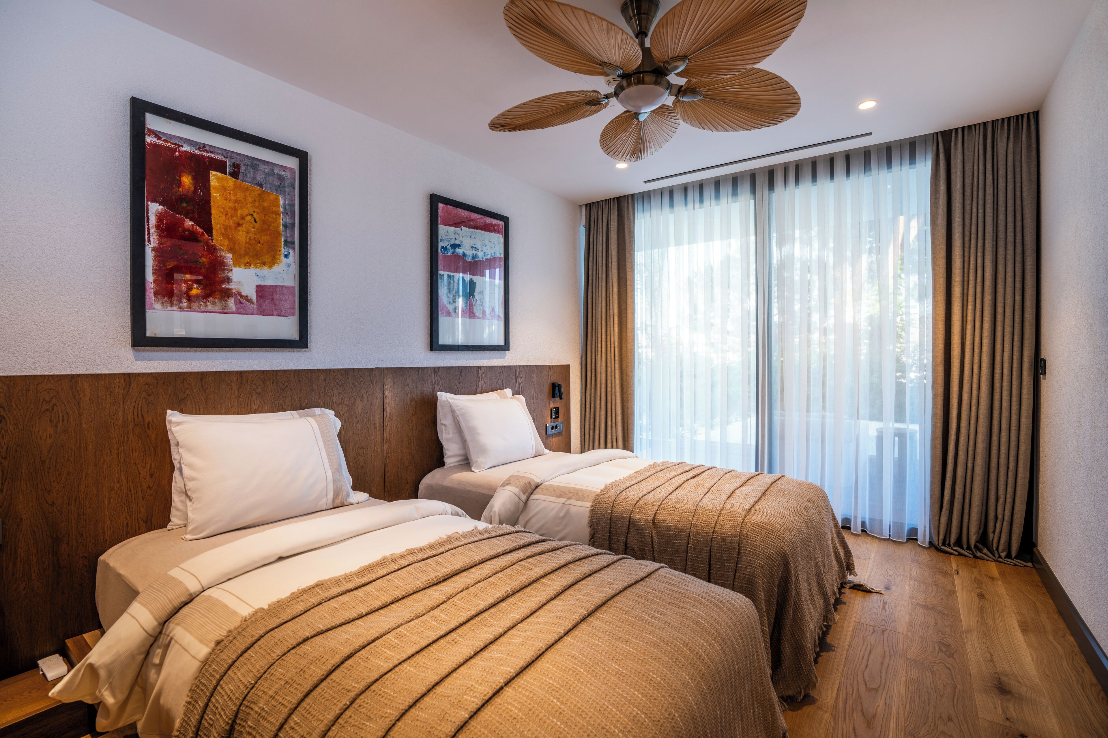 Habitación de hotel con dos camas, ventilador de techo, ventanas amplias con cortinas y obras de arte en las paredes.