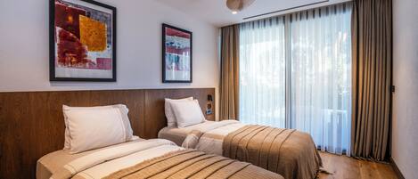 Habitación de hotel con dos camas, ventilador de techo, ventanas amplias con cortinas y obras de arte en las paredes.