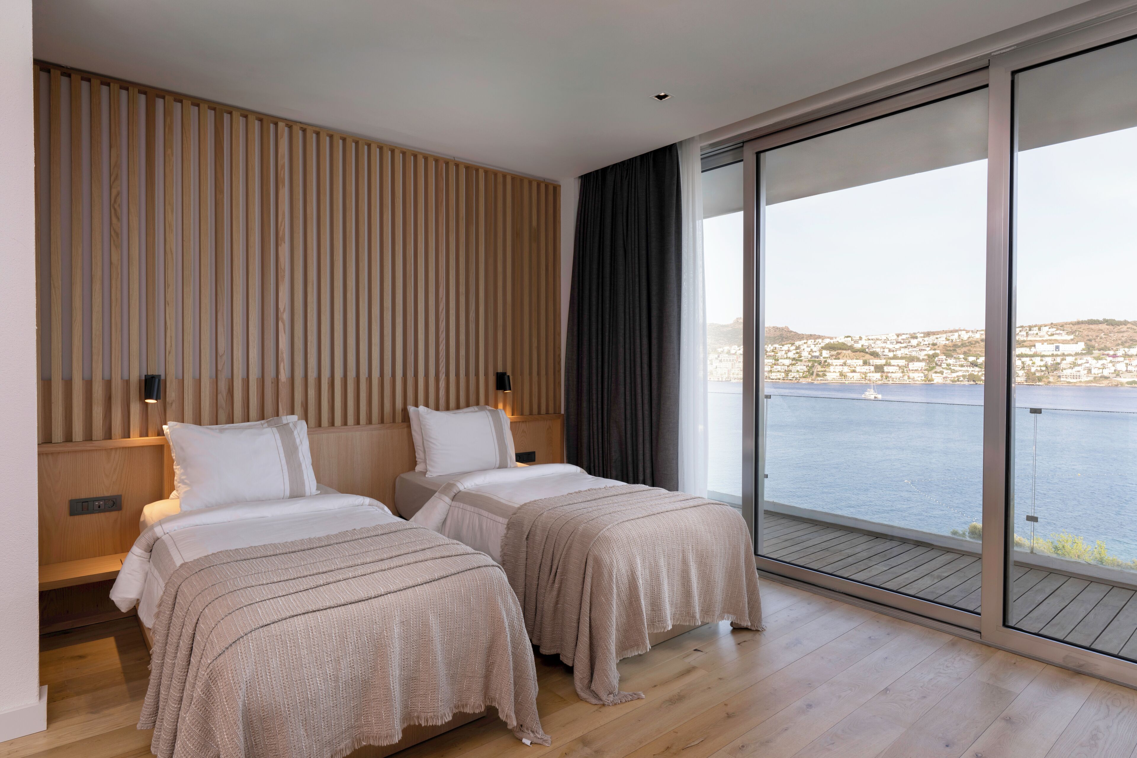 A modern hotel room with two beds, a wooden slat wall, a large sliding glass door to a balcony, and a view of a body of water and distant buildings.