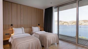 A modern hotel room with two beds, a wooden slat wall, a large sliding glass door to a balcony, and a view of a body of water and distant buildings.